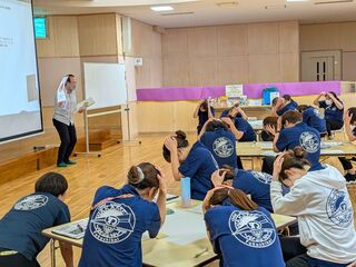 浦城こども園にて、講師がイラストの描かれたカードを掲げ、参加者たちが頭を抱えるポーズをとっている。