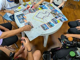 テーブルの上に、書き込みのある地図がある。数名でテーブルを囲んでいて、体育着の人や車いすに乗った人がいる。