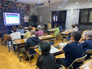 講師が壇上で話していて、参加者が話を聞いている様子。参加者には高齢者が多いが、子供もいる。