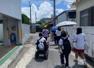昼間に、中学生から大人まで、7名ほどのグループで歩いて、住宅地をチェックしている様子。車いすに乗った人や、帽子をかぶった人、日傘をさした人がいる。